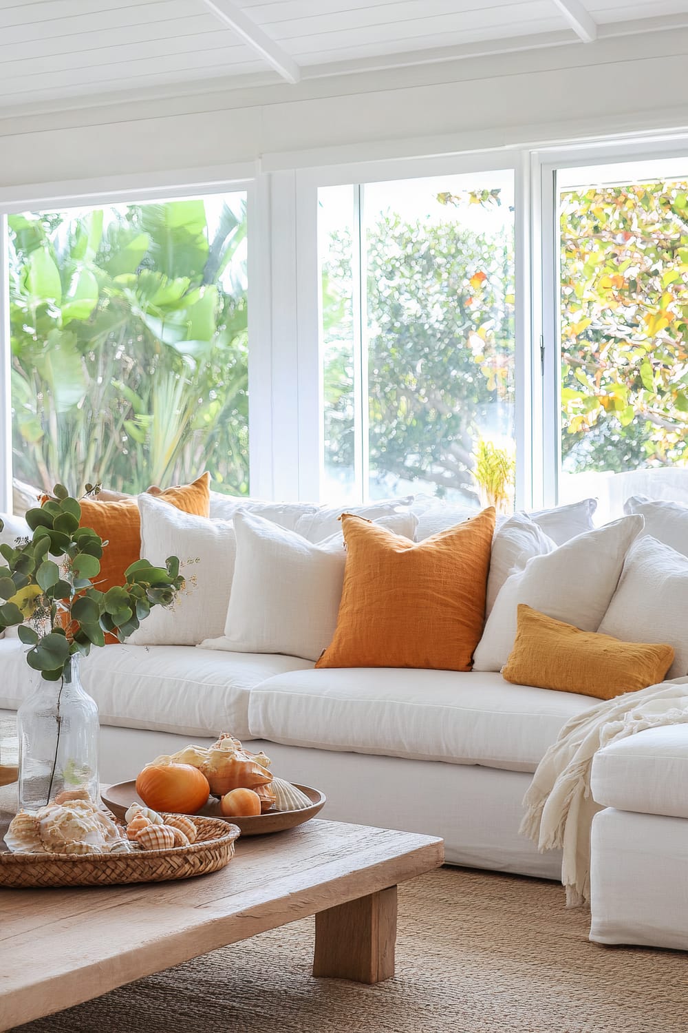 A bright living room with a white sectional sofa adorned with white, mustard yellow, and orange cushions. A light wooden coffee table holds a clear glass vase with green foliage, and wicker trays filled with seashells and decorative elements. Large windows in the background reveal lush green foliage outside.