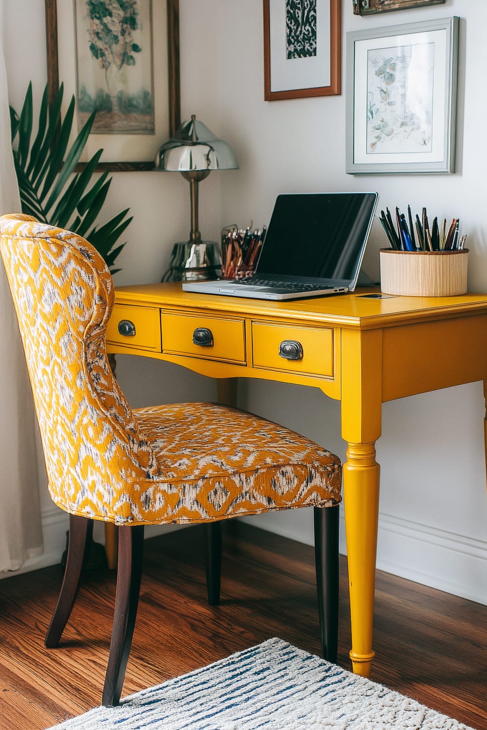 An inviting home office space featuring a yellow desk with vintage-style hardware, a laptop, and various pens in a circular organizer. The desk is paired with a cushioned chair displaying an intricate gold and white pattern. On top of the desk, a modern silver lamp is placed beside the laptop. Wall-hung artwork and leafy green plants add decorative touches to the setting. The floor is covered with a striped rug on a hardwood surface.