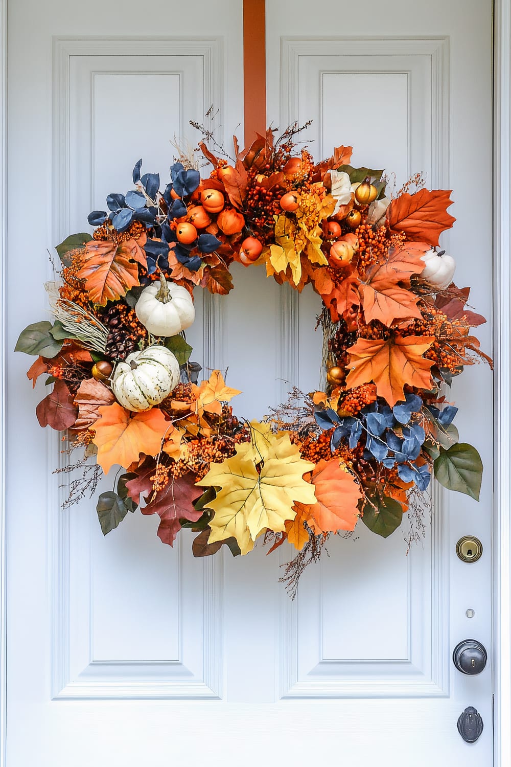 An autumnal wreath with a variety of decorative elements is hanging on a white front door. The wreath includes vibrant orange, red, yellow, and green leaves, faux mini pumpkins, both orange and white, and clusters of red berries. There are also smaller blue leaves and twigs incorporated throughout the design. The wreath is attached to the door with an orange ribbon, creating a festive and welcoming season-appropriate decoration.