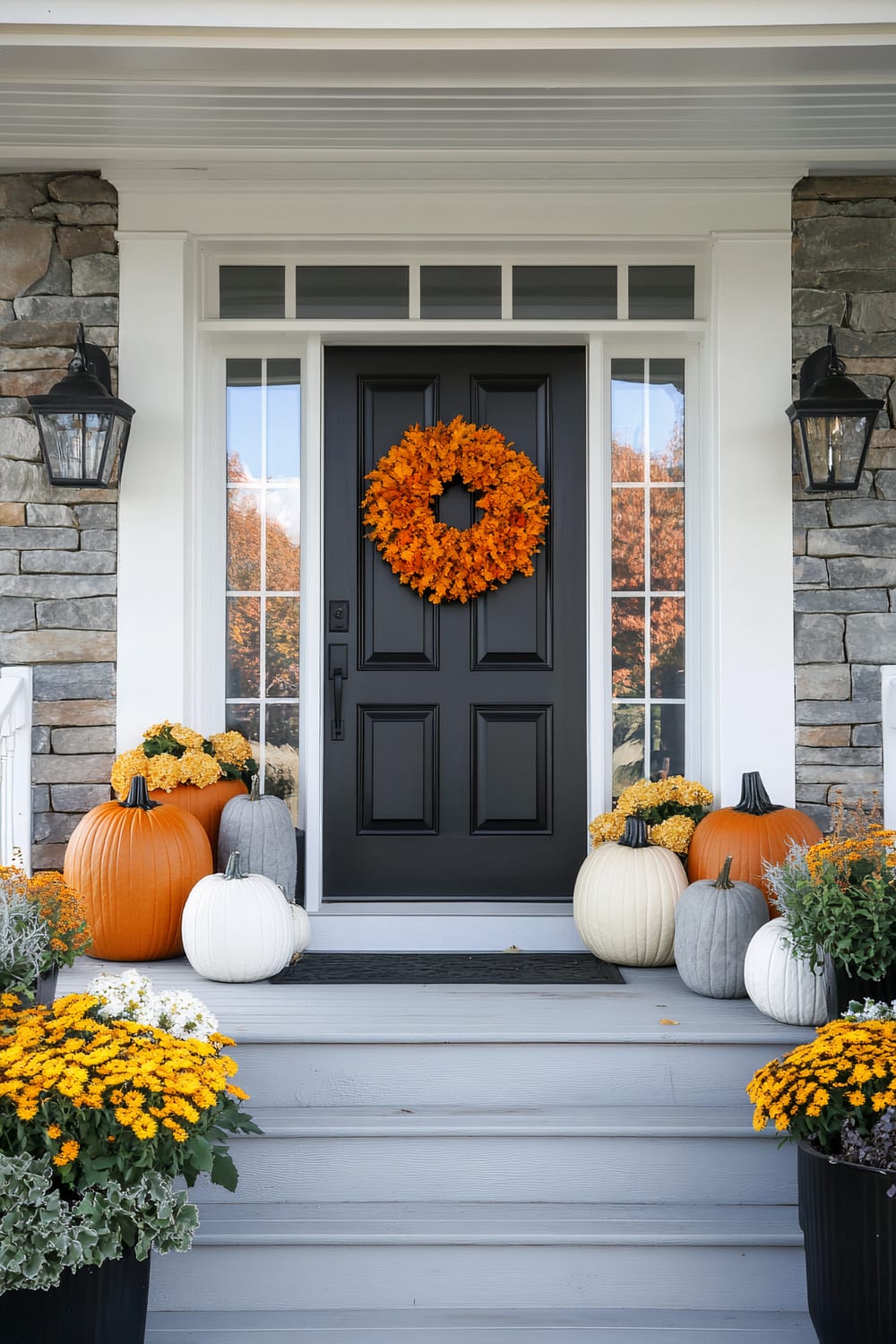 A front porch decorated for autumn. The door is black with an orange wreath hanging on it. Various pumpkins in orange, white, and grey are arranged on either side of the door, along with pots of yellow and white chrysanthemums. Stone walls flank the doorway, and black lantern-style light fixtures are mounted on them.