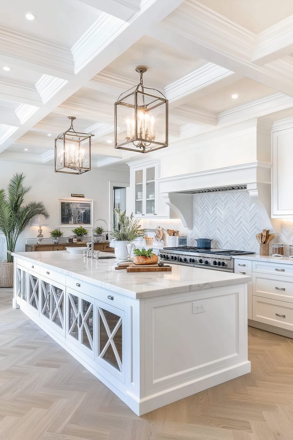 This image showcases a luxurious kitchen with a large marble-topped island that features cabinetry with glass-paneled doors. Two large, elegant lantern-style pendant lights with visible bulbs hang from a coffered ceiling, illuminating the space. The kitchen cabinets are white and there is a chevron-patterned backsplash above the stainless steel oven. Potted plants and fresh bread on a wooden cutting board adorn the countertops, adding a touch of nature and liveliness to the pristine kitchen. In the background, a potted palm and framed artwork enhance the decor.