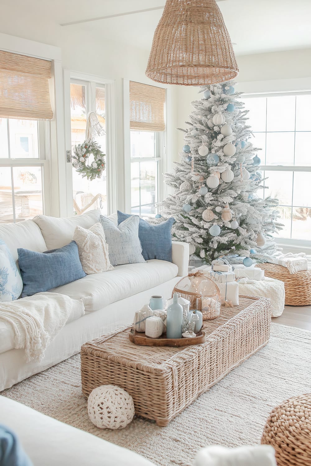 Bright coastal-themed living room featuring a large frosted Christmas tree adorned with blue and white ornaments. A white sofa with blue and white throw pillows sits to the left, with a rattan coffee table in front, displaying vases and decorative items. Large windows let in abundant natural light, adorned with wooden blinds and a festive wreath. The room is accented with a hanging wicker pendant light and additional woven baskets and decor.