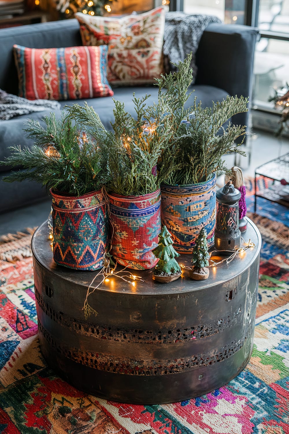 A colorful and eclectic living room scene featuring a round metal coffee table with perforated designs on its side. The table is decorated with woven baskets containing green pine branches adorned with string lights. There are also small decorative trees and lanterns on the table. Behind the table, a dark grey sofa is visible with multiple colorful, patterned throw pillows. The floor is covered with a vibrant multi-colored rug, adding to the bohemian aesthetic.