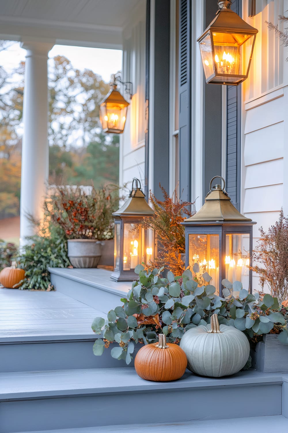 A well-decorated porch in autumn featuring white siding and blue shutters. Two large, metal lanterns with glowing candles are placed on the porch steps, surrounded by potted plants and decorative foliage. Two pumpkins, one orange and one pale green, are on the steps. Overhead, a gaslamp-style light adds a warm ambiance, while large white columns frame the porch.