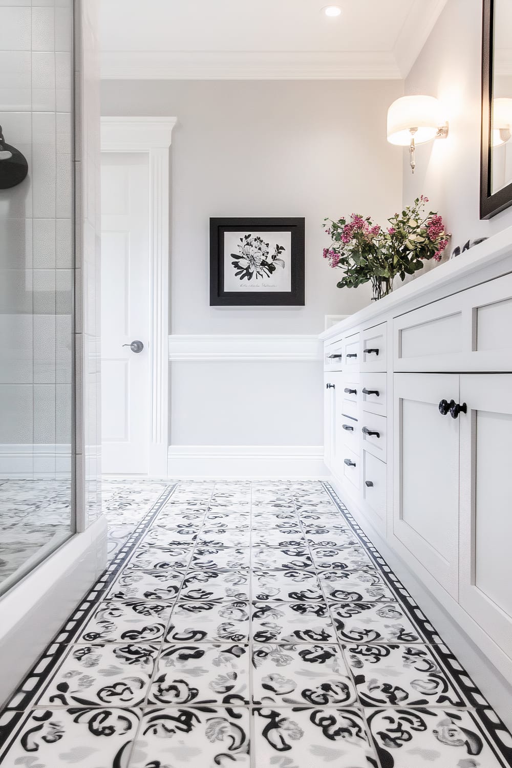 A chic bathroom featuring intricate black and white patterned floor tiles, complementing white cabinetry with black handles. The walls are painted a soft grey, adorned with a floral framed artwork and a potted plant with pink flowers. A wall-mounted light fixture illuminates the space.