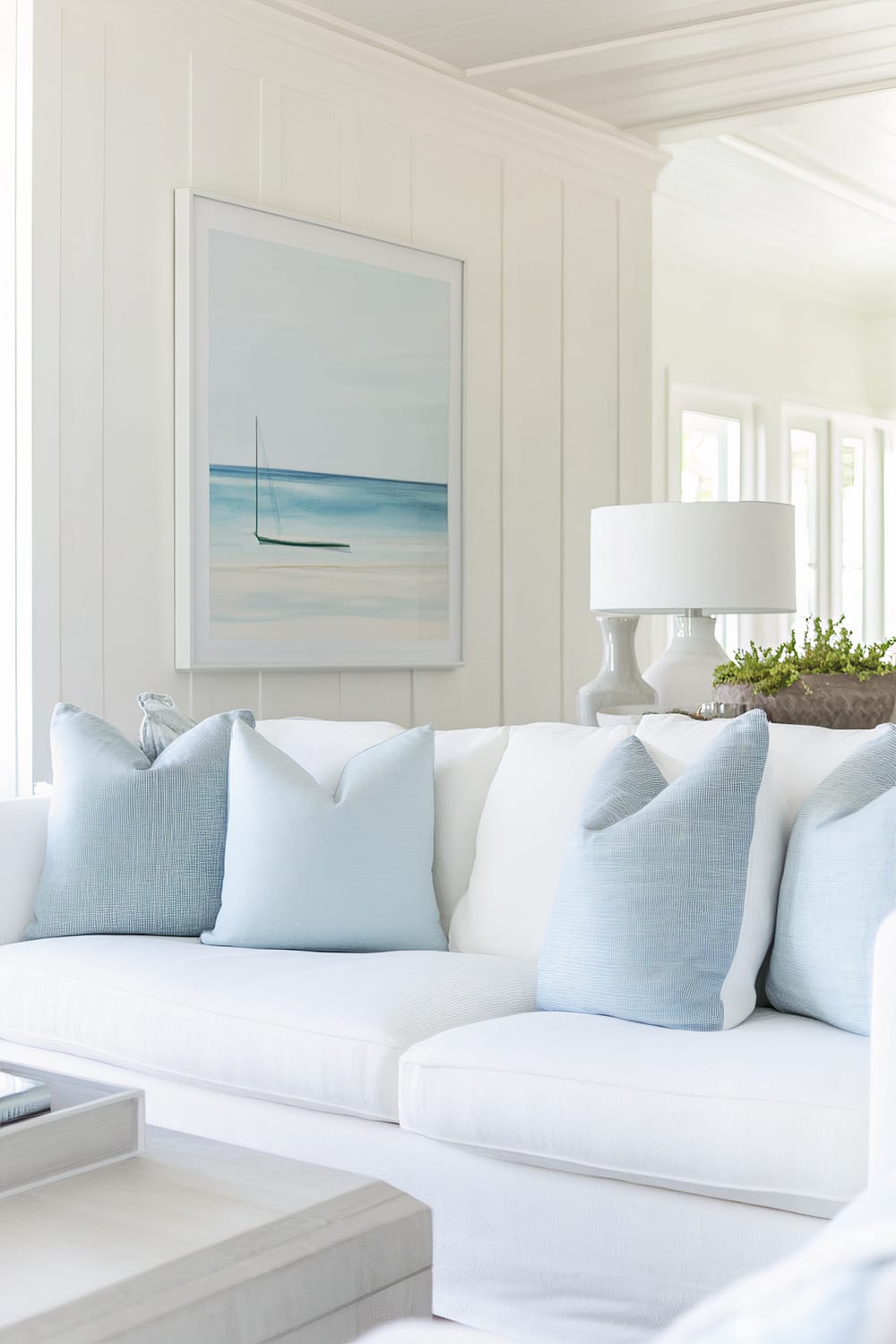 An elegant living room features a white couch adorned with light blue pillows, a serene ocean painting on a white-paneled wall above it, and a modern table lamp on a side table next to a green plant. The room is illuminated by natural light from tall windows.