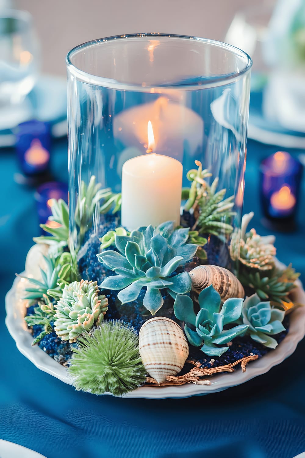 A striking table centerpiece featuring a large clear glass terrarium filled with vibrant blue and green succulents, nestled on a white ceramic plate with subtle wave patterns. The terrarium is surrounded by votive candles in navy holders against a deep teal tablecloth background, emitting a calming blue glow that accentuates the sea-themed decor, comprising seashells and miniature driftwood pieces.