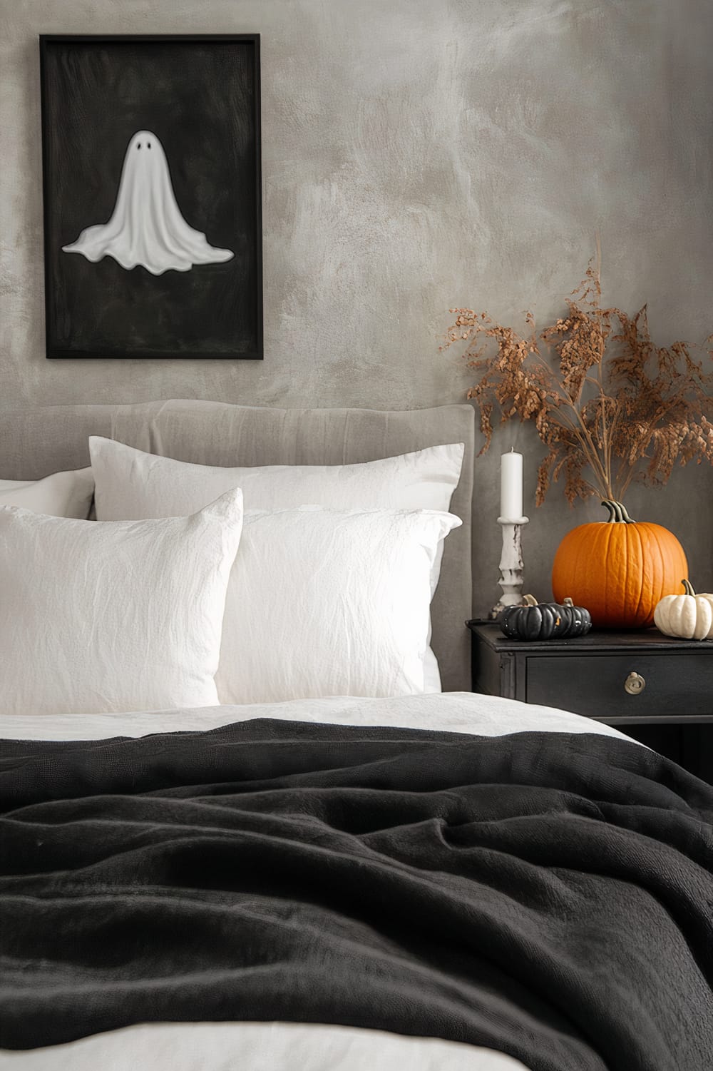 A bedroom featuring a minimalist Halloween theme, with a bed covered in white pillows and bedding, accented by a black throw blanket. A ghost painting hangs on a gray-toned wall above the bed. The bedside table holds a pumpkin, a white candle, and dried foliage.