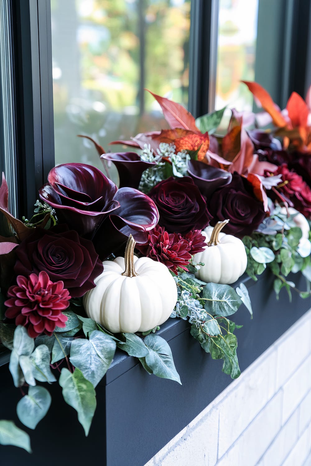 A window box filled with a rich arrangement of deep burgundy flowers, including calla lilies and roses, complemented by white ornamental pumpkins and varied green foliage. The window itself is framed in black, and the planter box is set against a brick wall exterior.