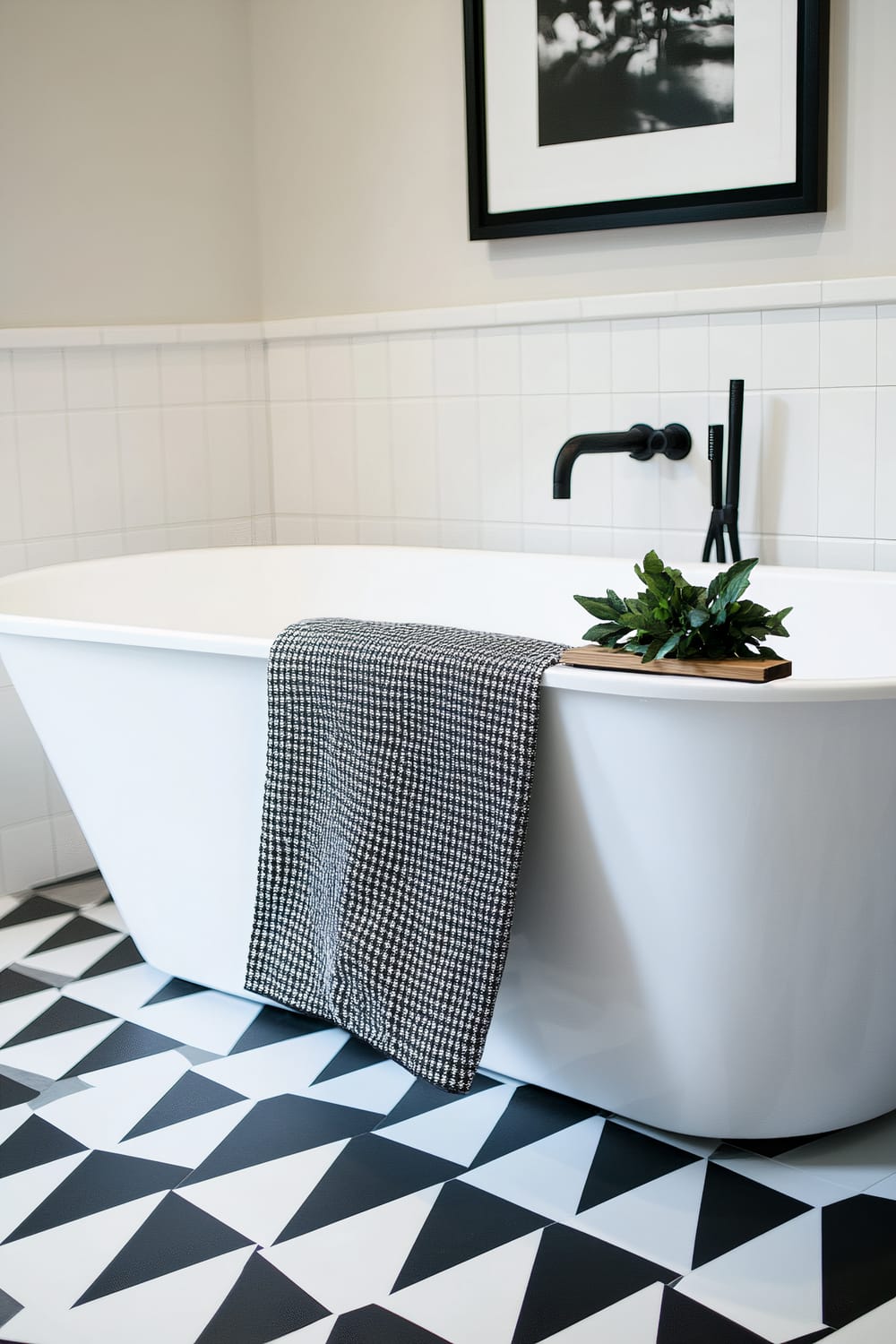 A modern bathroom features a white freestanding bathtub as the centerpiece. Draped over the edge of the tub is a black and white patterned towel. A wooden bath caddy resting on the tub holds a green plant. The floor is covered in black and white geometric tiles arranged in a triangular pattern. The wall above the bathtub has rectangular white tiles, and a black-framed monochrome photograph hangs on the wall.