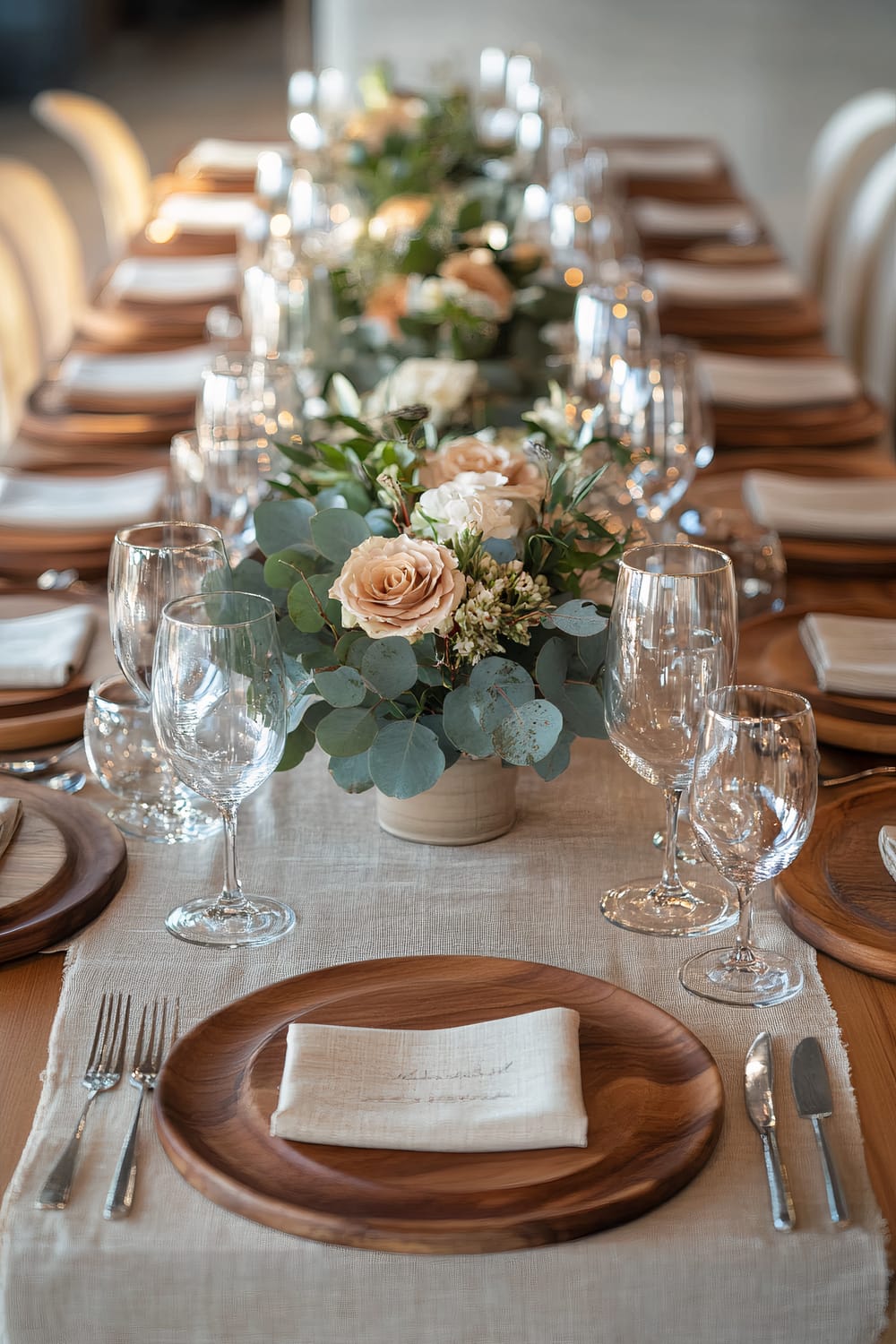 A long dining table is elegantly set for a Thanksgiving meal. The table features a beige linen runner, natural wood chargers, and neatly folded cloth napkins. In the center are muted floral arrangements with eucalyptus leaves and light-colored roses. Clear wine glasses and silver cutlery complete the setting, all illuminated by warm, directional lighting creating a cozy atmosphere.