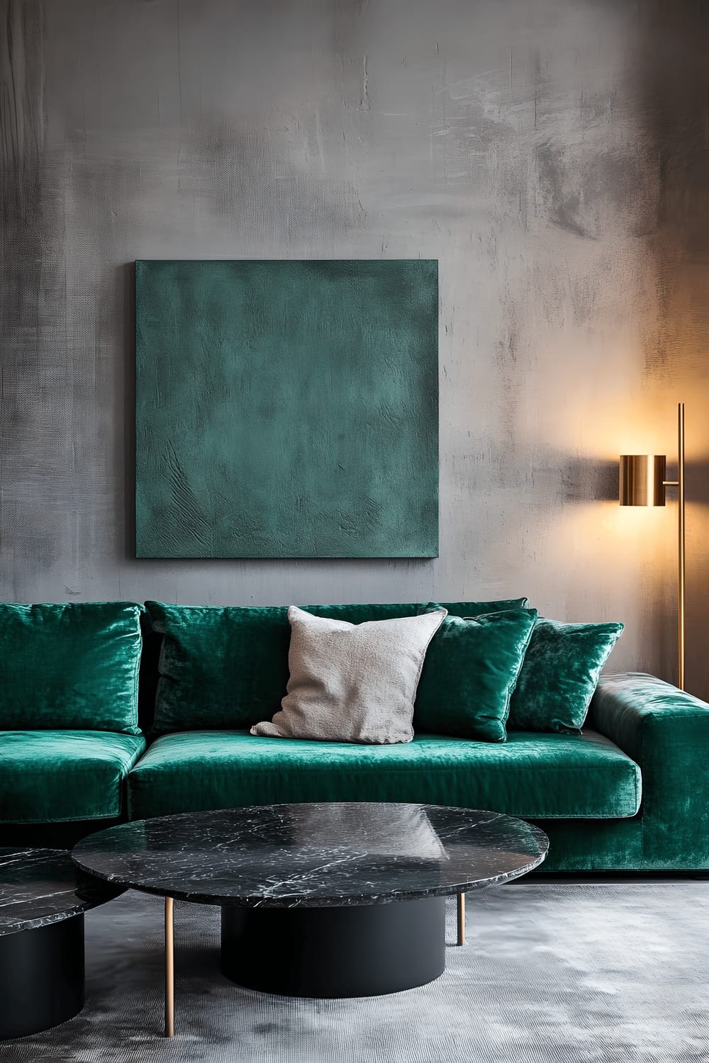 A warm, inviting lounge area featuring a large emerald green velvet sectional sofa resting against a striking charcoal gray concrete accent wall. In front of the sofa, a matte black marble coffee table sits on a plush rug with complementary cobalt blue metal side tables flanking the sofa. Brassy gold floor lamps with leaf motifs are strategically placed to cast a soft, warm light on the sofa and the coffee table. The wall is minimalistically decorated with a single piece of wall art keeping the look clean and clutter-free.