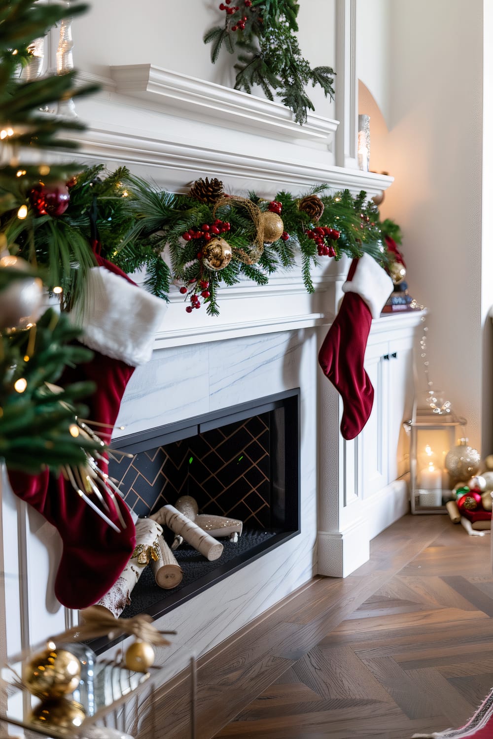 An elegantly decorated fireplace adorned with festive Christmas decor. It features green garlands embellished with gold and red baubles, pinecones, and berry branches. Two red and white stockings are hung from the mantle. A Christmas tree with lights and ornaments is partially visible on the left. A lantern with a candle and additional Christmas ornaments are placed on the floor near the fireplace.