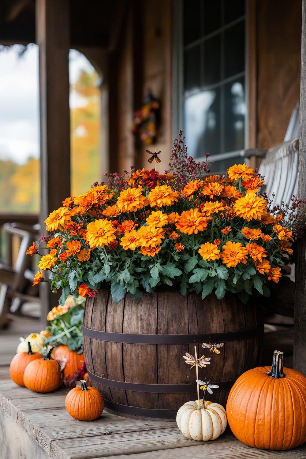 An autumn-themed porch scene features a rustic wooden barrel filled with vibrant orange and yellow flowers. Surrounding the barrel are small and medium-sized pumpkins, enhancing the fall ambiance. A dragonfly ornament is attached to one of the pumpkins, adding a whimsical touch. The porch includes wooden railings and a chair visible in the background.