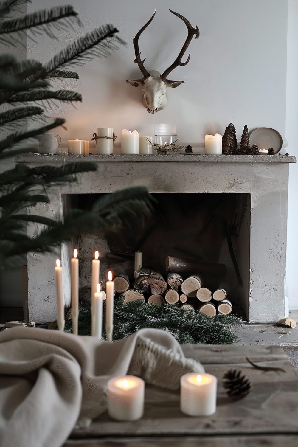 An intimately styled interior featuring a rustic fireplace adorned with candles of varying sizes and pine cones on the mantel. Above the mantel is a mounted deer skull with antlers. Logs are neatly stacked inside the fireplace, with evergreen branches and more lit candles placed in front on a wooden surface. A beige blanket is draped over the wooden surface, adding a cozy element to the setting.