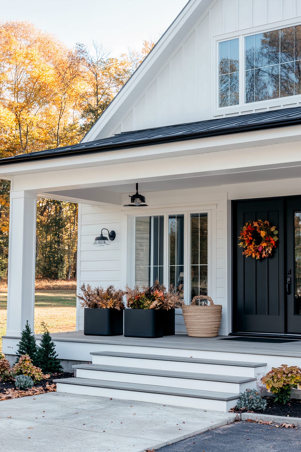 An inviting front porch of a white house with clean architectural lines, a black door decorated with an autumn wreath, and three black planters with fall leaves. A wicker basket sits beside the door. The porch features white steps leading down to a concrete pathway. Trees with colorful autumn foliage are visible in the background.