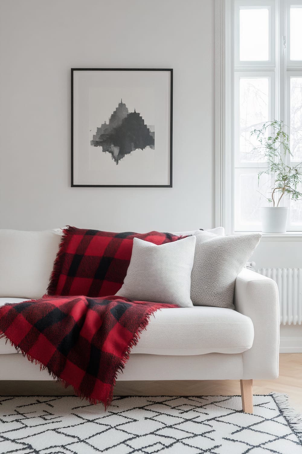 An interior living space featuring a white sofa with a red and black plaid blanket draped over it. The sofa also has two gray cushions. Above the sofa hangs a black-and-white abstract painting in a black frame. To the right is a large window letting in natural light with a potted plant on the window sill. Beneath the sofa is a white area rug with black geometric patterns.