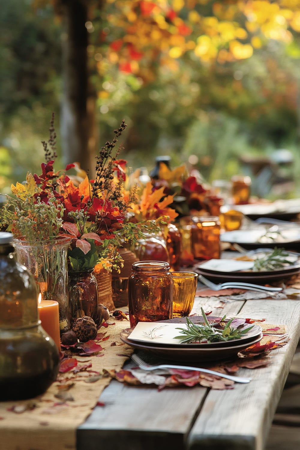 An outdoor dining table set for a fall-themed meal. The table is decorated with amber glass jars holding candles and autumn floral arrangements with vibrant leaves in red, orange, and yellow. A burlap table runner and scattered autumn leaves add to the seasonal decor. Place settings include dark brown plates with sprigs of greenery, and polished silverware. The background shows blurry autumn foliage in golden hues.