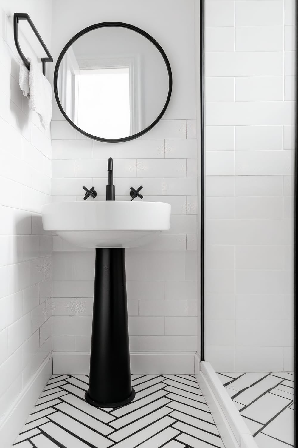 Compact bathroom featuring a sleek white pedestal sink with a matte black base, complemented by a round black-framed mirror mounted above. The wall behind the sink is tiled in white subway tiles, and the floor showcases a white herringbone tile pattern with black grout. A wall-mounted, minimalist towel rack with a white hand towel is visible to the left, and a shower or partition with a glass panel is seen to the right.