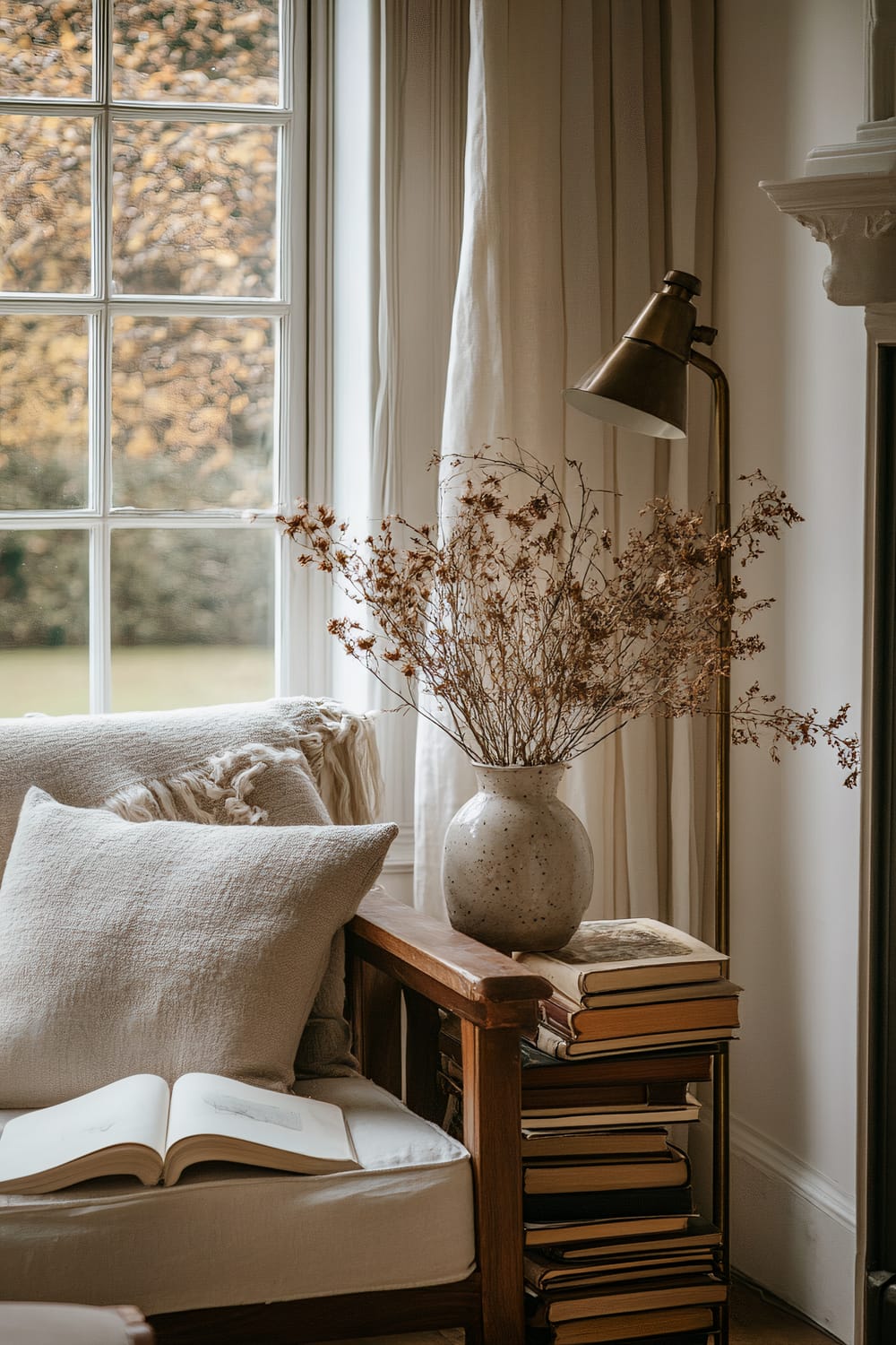 Sunlit cozy corner with a soft armchair next to a large window, adorned with beige cushions and a knitted throw. A wooden side table holds a rustic ceramic vase with dried flowers and a stack of books, complemented by a brass floor lamp and thick, cream-colored curtains. Outside, autumn foliage can be seen through the window.