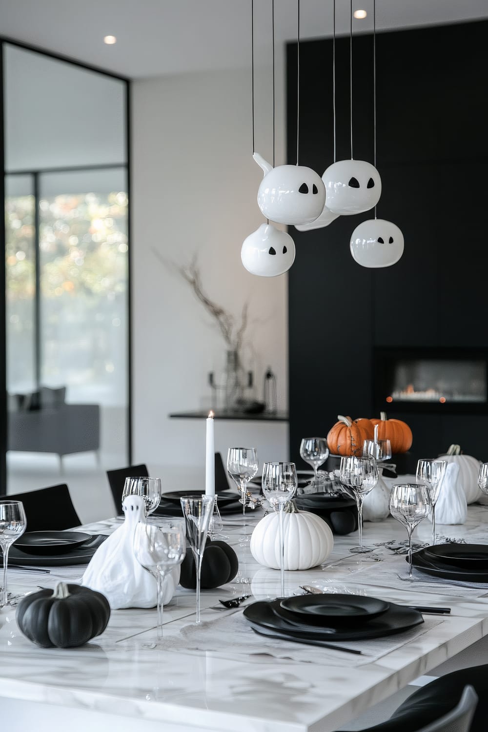 A modern dining room elegantly decorated for Halloween. The marble table is adorned with black plates set on gray and white table runners, surrounded by clear wine glasses and silverware. Small decorative pumpkins in black, white, and orange, along with white ghost figurines, are scattered across the table. Above the table, suspended from the ceiling, are unique ghost-shaped pendant lights. The room features a minimalistic aesthetic with large glass windows, a fireplace in the background, and a black accent wall.