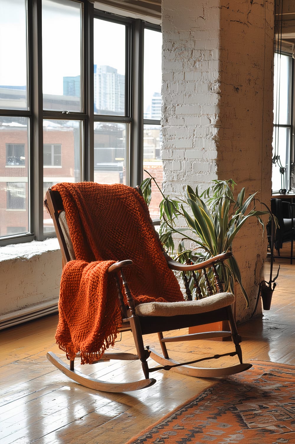 An interior scene features a wooden rocking chair with a chunky orange blanket draped over it. The chair is placed near a large window with a view of urban buildings outside. Next to the chair is a tall green plant. The room has a rustic industrial vibe with exposed brick walls and wooden floors, complemented by an orange patterned rug.