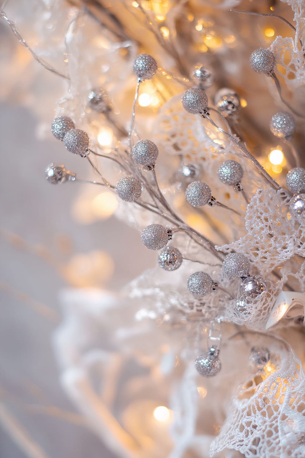 A closeup of a white lace wreath decorated with small silver berries and glowing fairy lights. The texture of the lace and the glittering surface of the berries are highlighted by the soft lighting, creating a timeless holiday charm. The background is muted and blurred.