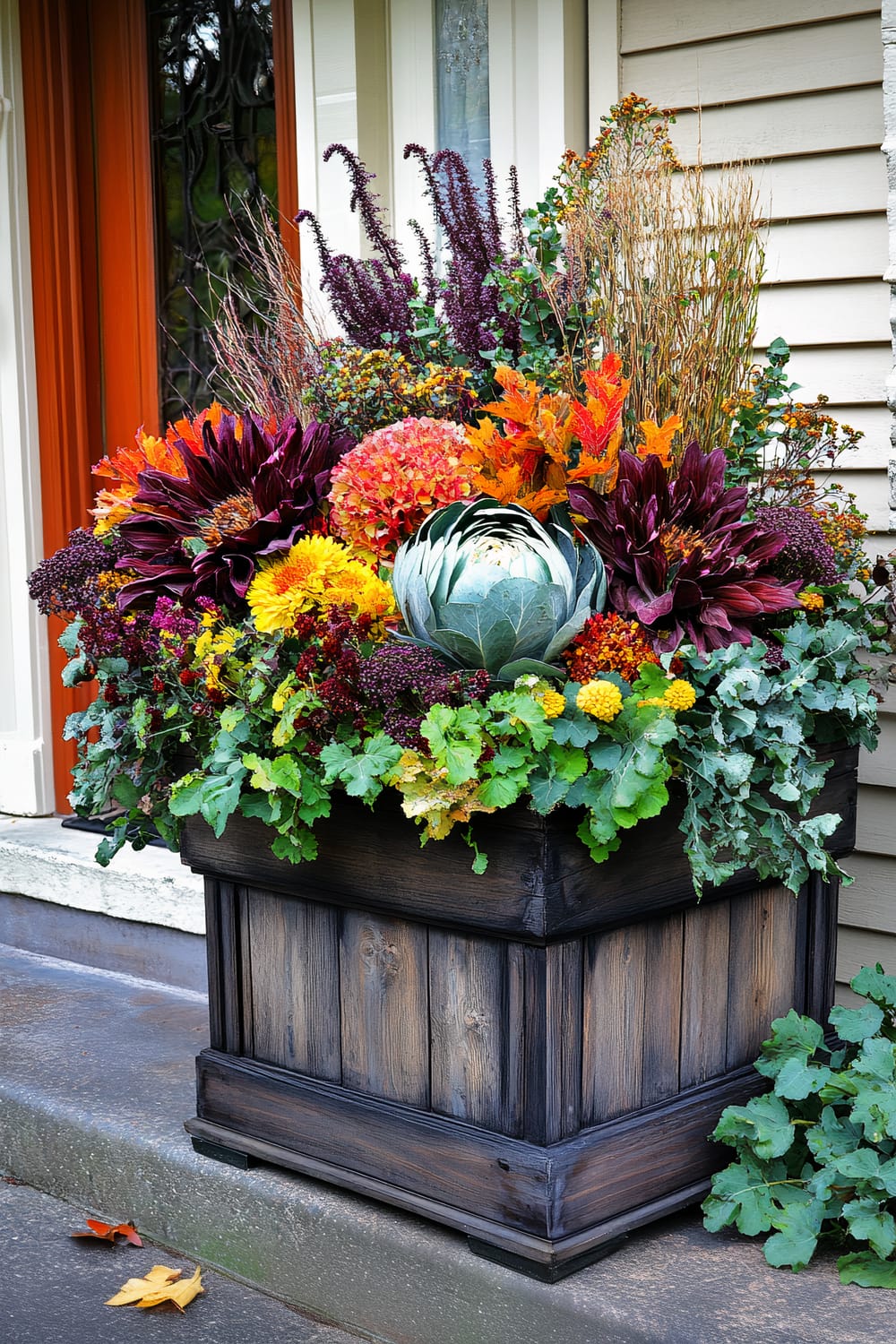 A vibrantly colorful autumn flower arrangement sits in a rustic wooden planter box situated at the entrance of a home. The planter box is weathered with a dark stain, blending seamlessly with the natural hues of the flowers. The arrangement boasts a striking mix of fall foliage and flowers, including deep burgundy, bright orange, and rich yellow blooms, along with green leafy vegetables such as a cabbage and kale. The backdrop features a beige exterior wall with vertical siding and an adjacent reddish-orange door frame, enhancing the fall-themed aesthetic.