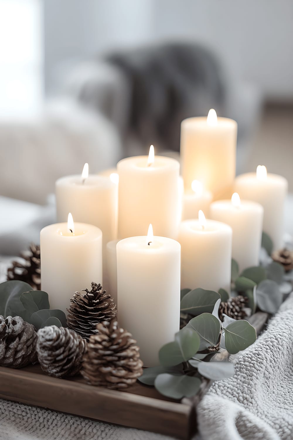 A Scandinavian-inspired dining table centerpiece with a collection of white and pastel colored candles of varying heights arranged in simple glass holders. The candles sit on a light wooden tray, adorned with sprigs of eucalyptus and pinecones. Anchoring the centerpiece is a soft gray wool runner. The backdrop reveals a minimalist table setting, illustrating an overall vibe of comfort and simplicity.