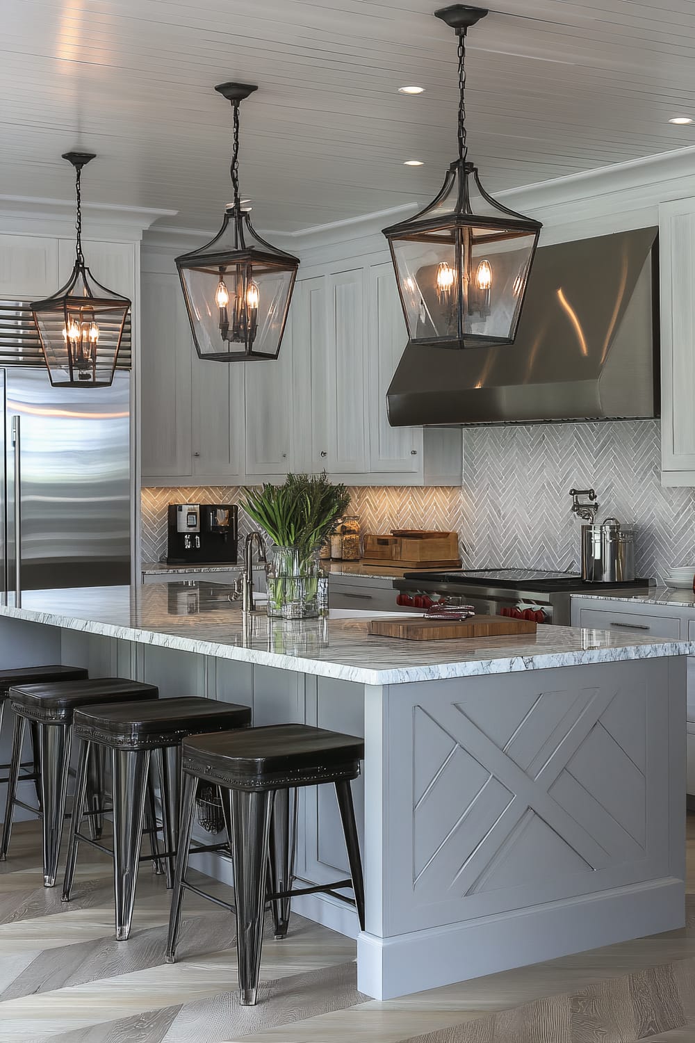 An elegant kitchen featuring a central island with a marble countertop and four black metal stools. Above the island, there are three large lantern-style pendant lights hanging from the ceiling. The kitchen cabinetry is painted in light grey with a stainless steel refrigerator and a large stove range with a metal hood. A herringbone-patterned backsplash in light grey and white hues adds texture to the space. A vase with fresh greens sits on the island, adding a touch of nature.