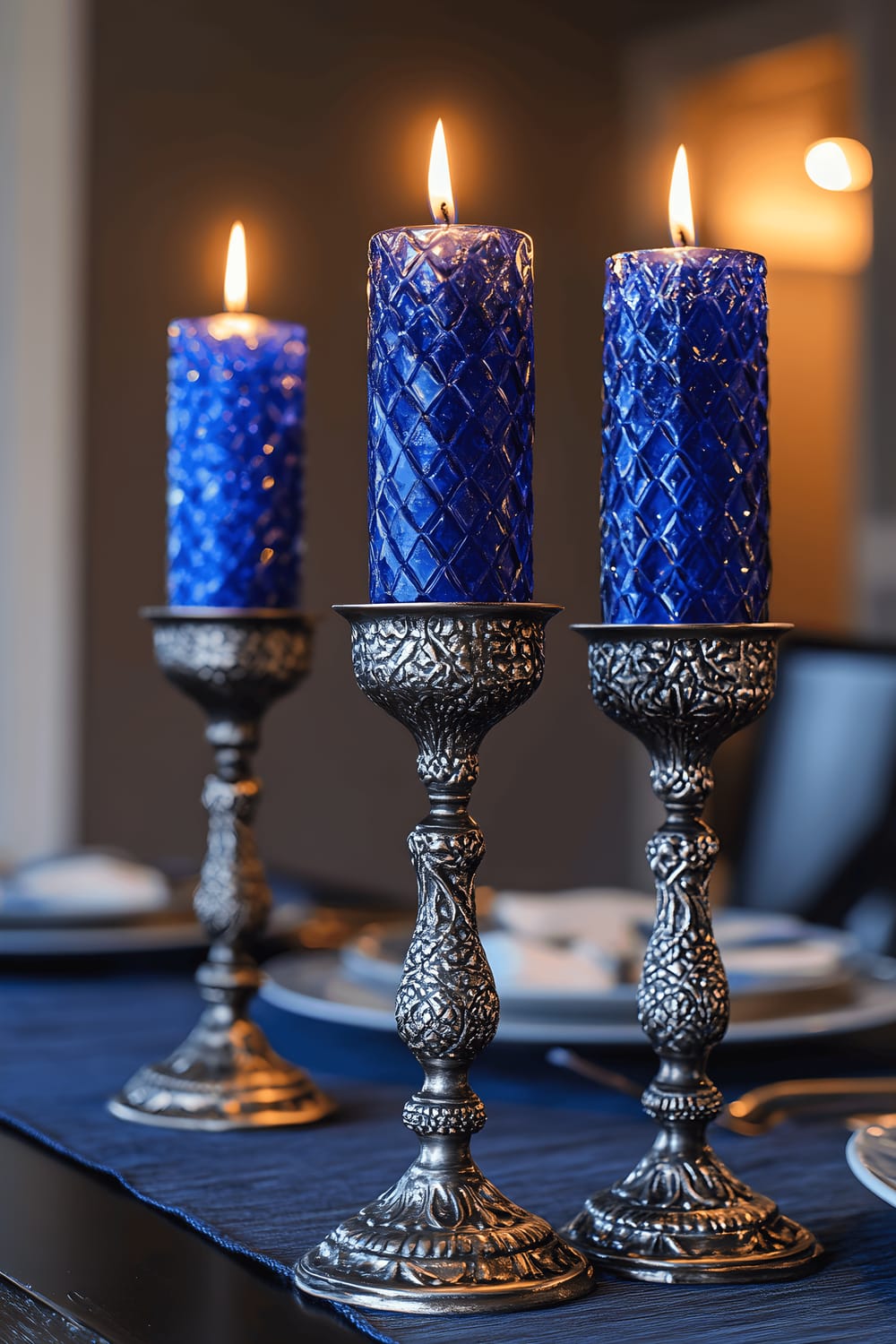 A captivating dining room scene showcasing three pewter candle holders, each holding a cobalt blue crystal candle, center-placed on a reclaimed wooden table with a charcoal gray runner. These are under dramatic overhead lighting enhancing the deep blue and metallic tones. The room features dark walls and minimalist decor.