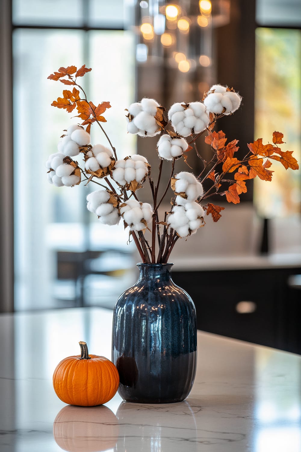 A dark, glossy vase with a bouquet of white cotton stems and autumn leaves is placed on a marble countertop. Next to the vase is a small, round pumpkin. The background shows a blurred view of a modern, well-lit interior, featuring large windows and contemporary light fixtures.