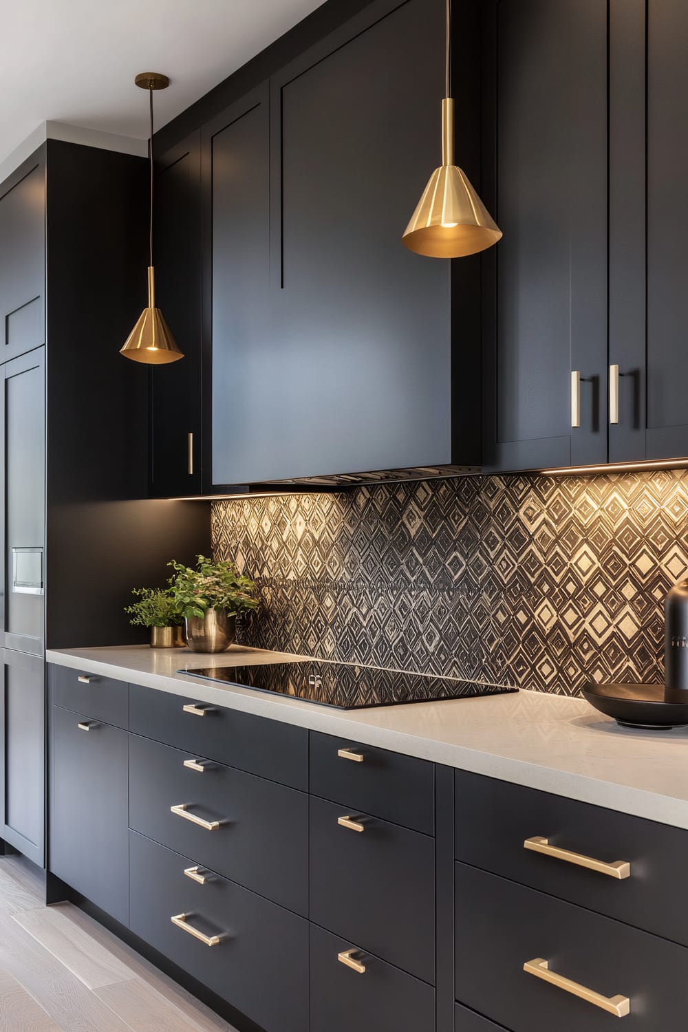 This modern kitchen features sleek black cabinetry with matte gold handles and fixtures, highlighted by a striking black and white geometric-patterned backsplash. Two pendant lights with gold cone-shaped shades hang above a white quartz countertop that houses an induction cooktop. A small brass pot with greenery adds a touch of natural color to the elegant, sophisticated space.