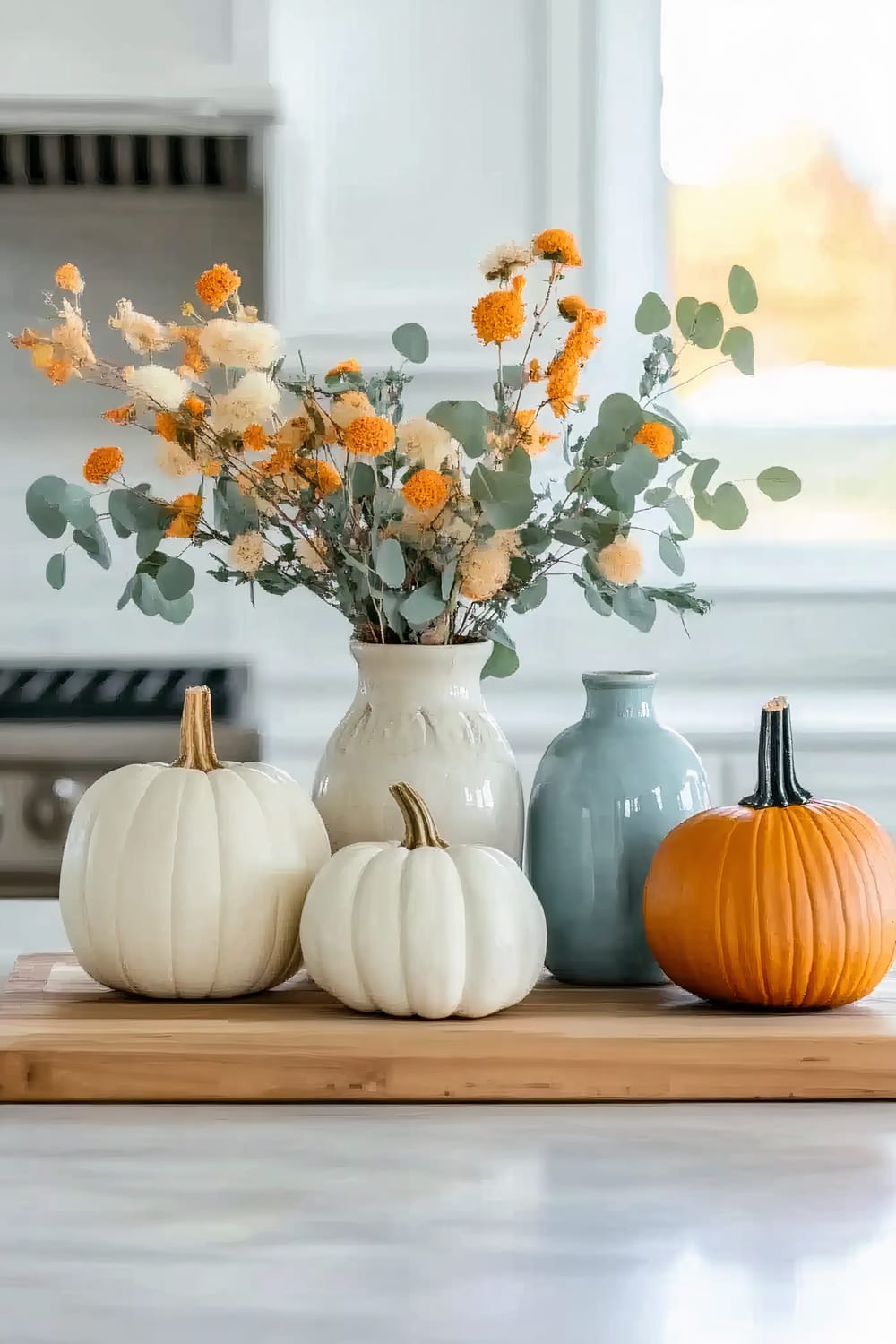 A kitchen scene featuring a decorated wooden cutting board with autumn elements. Three pumpkins—two white and one orange—are placed alongside a cream ceramic vase filled with orange and white dried flowers, and a smaller blue ceramic vase.