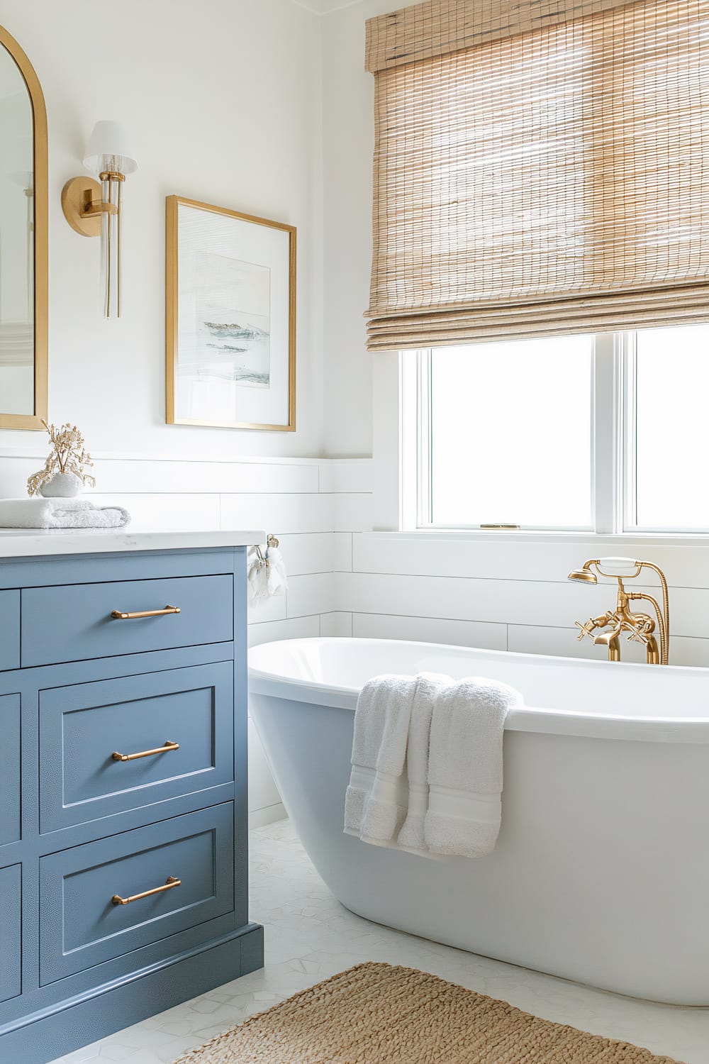 A sophisticated bathroom features a freestanding white bathtub with gold fixtures, positioned by a window framed by woven wooden blinds. Adjacent to the bathtub is a blue vanity with gold handles and a marble countertop, adorned with neatly folded white towels and a decorative object. Above the vanity hangs an oval mirror with a gold frame, complemented by a gold wall sconce with a white shade. A piece of framed minimalist artwork is mounted on the wall beside the mirror. The floor is covered with patterned tiles, and a woven jute rug adds texture to the space.