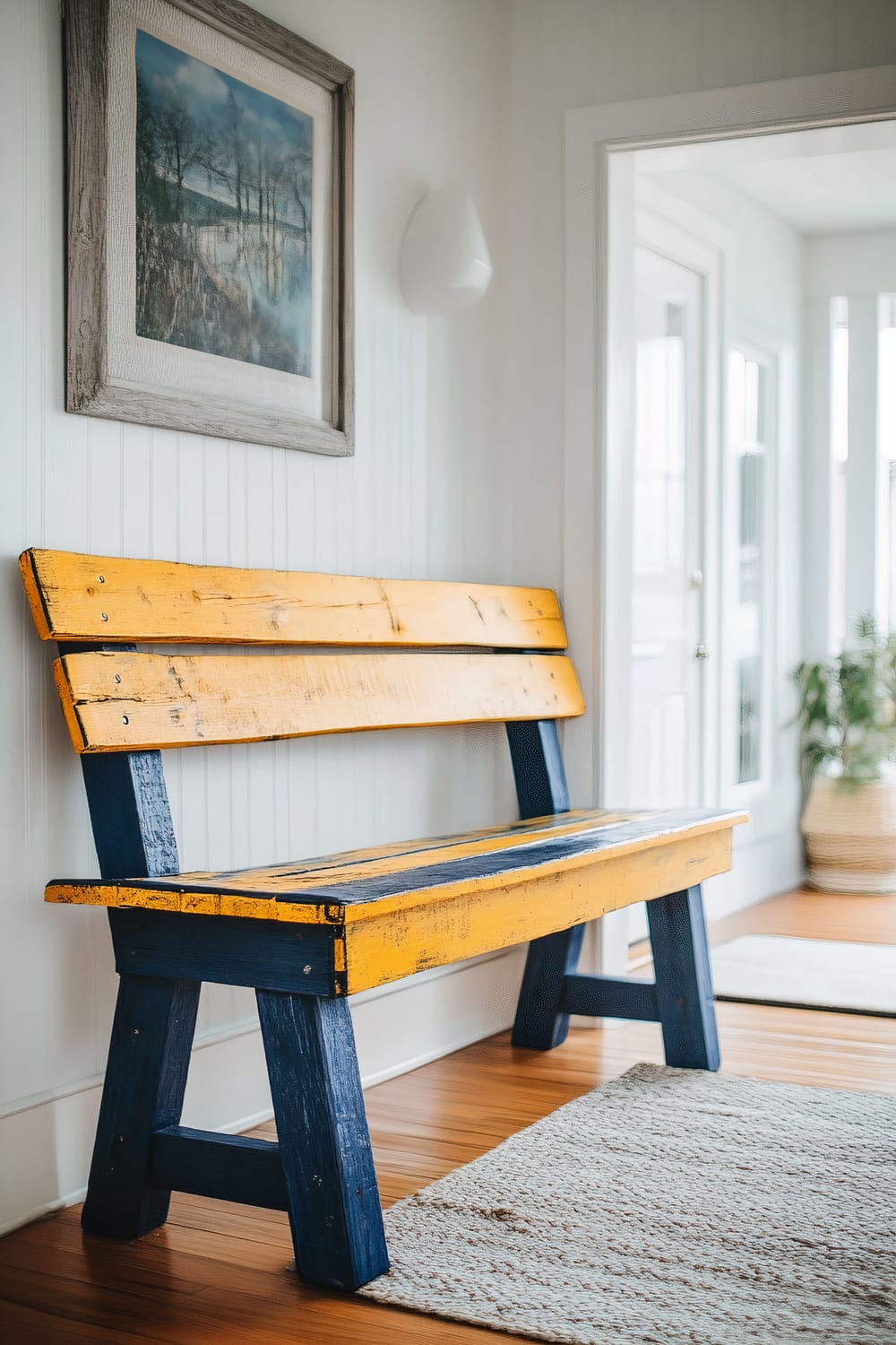 A yellow and navy blue bench made from repurposed wooden pallets placed in a white-walled entryway. The bench is rustic in design, and bright lighting emphasizes its bold colors. A white woven rug lies on the wooden floor, and a framed landscape painting hangs above the bench on the wall.