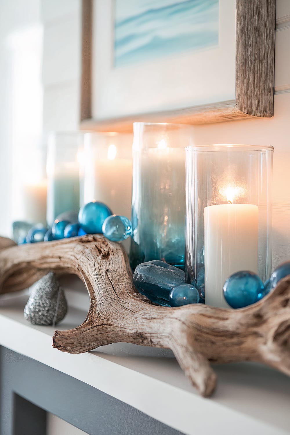 A decorative display featuring white pillar candles in glass cylinders, seafoam blue glass orbs, and a piece of driftwood on a shelf. The background includes a framed wall art with a hint of oceanic scenery.