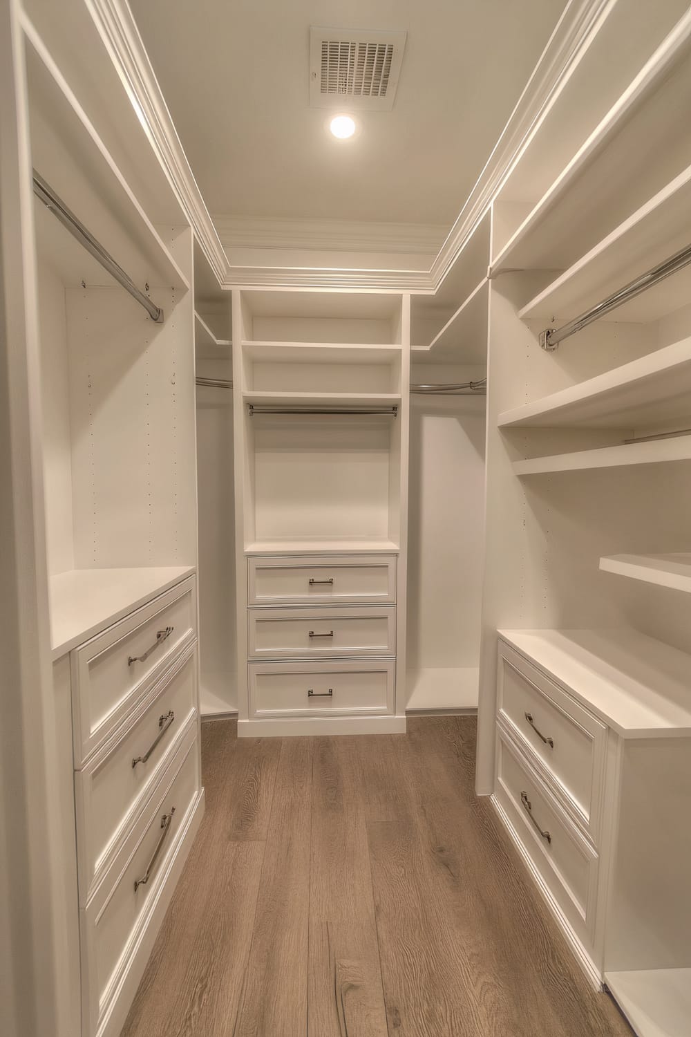 A compact walk-in closet with white built-in cabinets, drawers, and shelves, featuring chrome handles and a light oak wood floor.