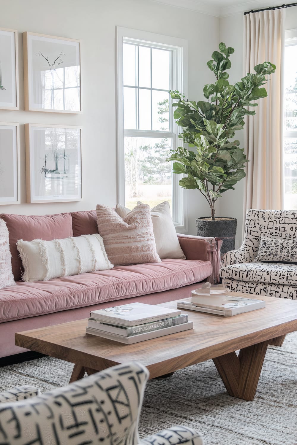 A chic living room features a pink velvet sofa adorned with various textured pillows. In front of the sofa, there's a wooden coffee table with sculptural legs, holding books and decorative trays. Two abstract-patterned armchairs in cream and black tones flank the coffee table. A set of six minimalist art prints hangs on the wall above the sofa. Large windows fill the room with natural light, and a tall potted plant stands near one of the windows, adding a touch of greenery.