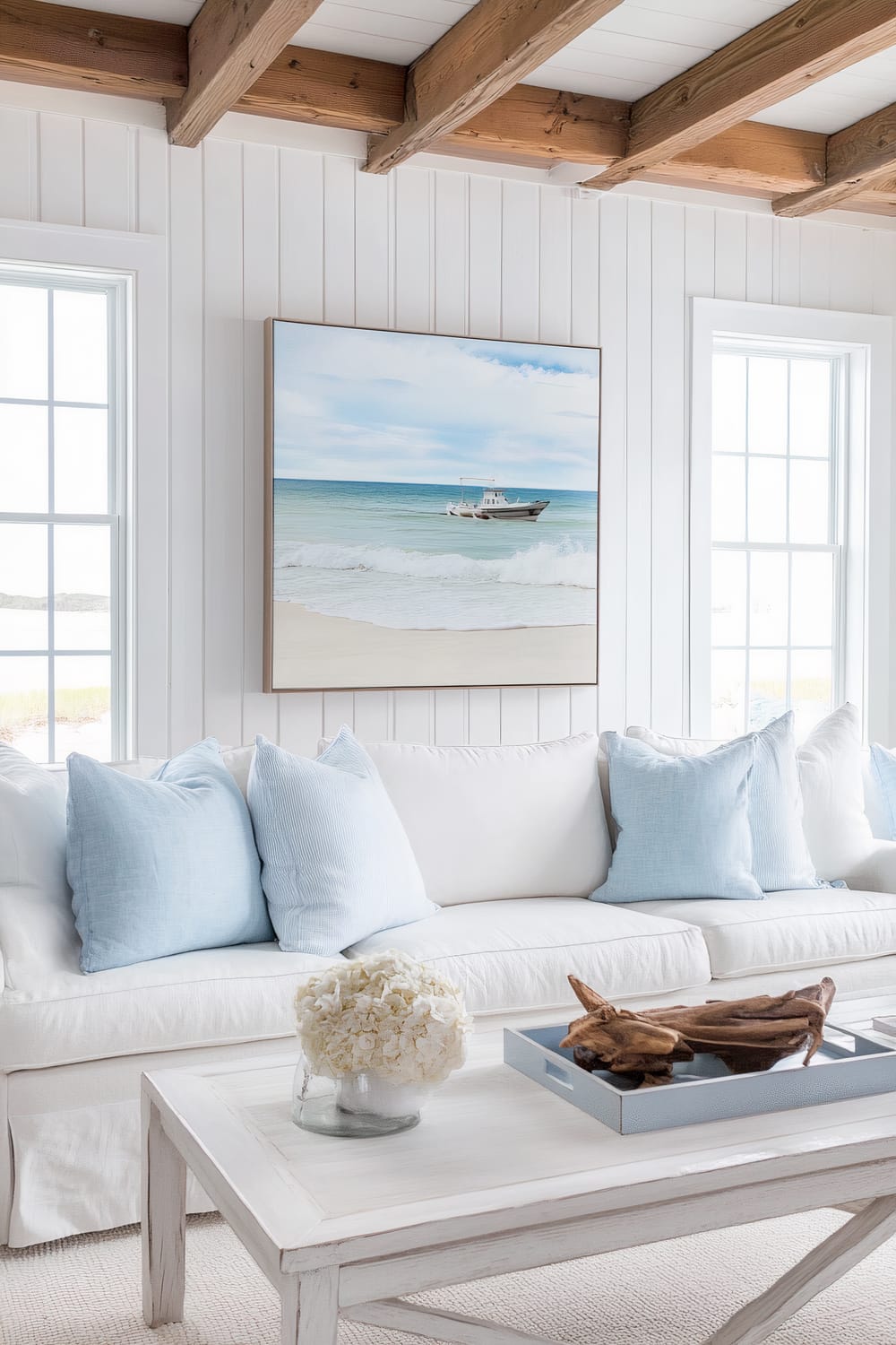 A bright, coastal-themed living room features a white couch adorned with light blue pillows. Above the couch, a large painting of a serene beach scene with a boat on tranquil waters hangs on a wall lined with white shiplap. The room is accentuated by large windows that allow natural light to flood the space. A simple white wooden coffee table in front of the couch holds a glass vase filled with white flowers and a blue tray with pieces of driftwood. Exposed wooden beams adorn the ceiling, adding a touch of rustic charm.