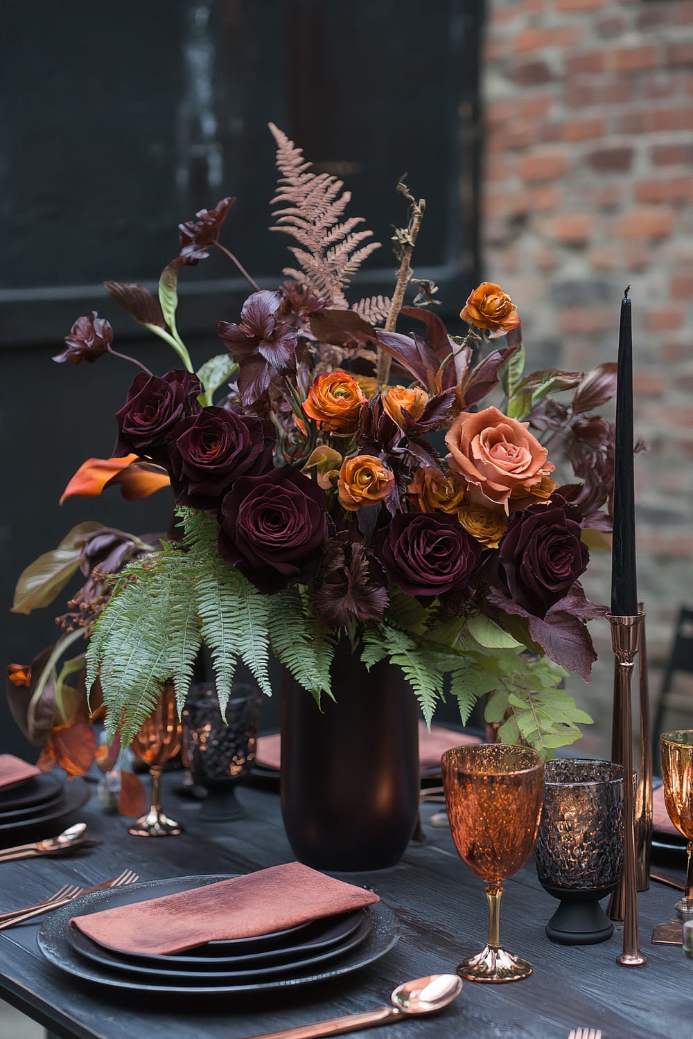 A dark, elegant table setting featuring a large floral arrangement of deep burgundy and orange flowers, green foliage, and maroon leaves in a black vase. The table is set with black plates, peach-colored napkins, and gold flatware. Amber and black goblets stand next to black candles in slim holders.