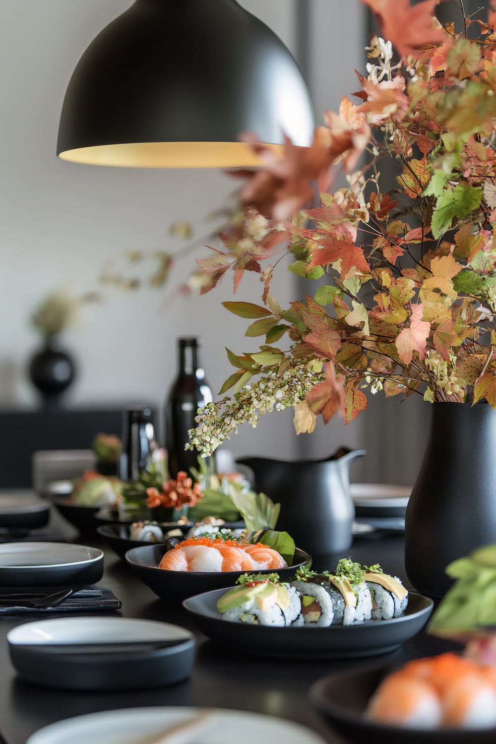 A modern dining table setup featuring plates of sushi, including turkey sushi rolls, avocado nigiri, and seasonal vegetable tempura, with a minimalist floral arrangement of autumn leaves and metallic accents at the center. Elegant sake sets and vintage-inspired pitchers are also present. The scene is illuminated by ambient lighting from a large black pendant lamp, creating a sophisticated atmosphere.