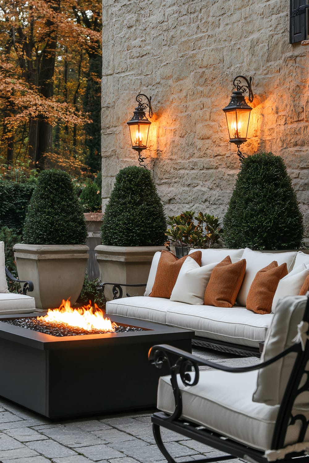 An outdoor seating area featuring beige cushioned sofas and a chair with black iron frames. The sofas are adorned with white and orange throw pillows. At the center is a black rectangular fire pit with glowing flames. Behind the seating area, a beige stone wall houses two vintage-style lanterns, lit and casting a warm glow. The space is surrounded by potted, manicured shrubs and trees with autumn foliage.