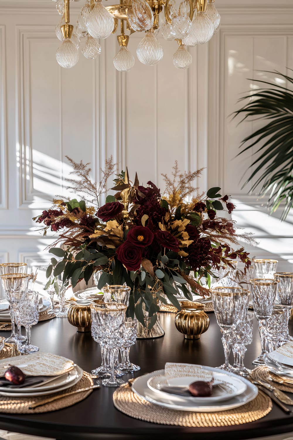 A luxurious dining table is beautifully set with crystal glasses and fine china adorned with gold accents. The centerpiece is a lush floral arrangement featuring deep red and burgundy flowers complemented by golden leaves and delicate foliage. Surrounding the arrangement are golden pumpkins and votive candles. A sophisticated chandelier with glass pendants hangs above, and the room's elegant white, paneled walls and bright ambiance add to the opulent feel.