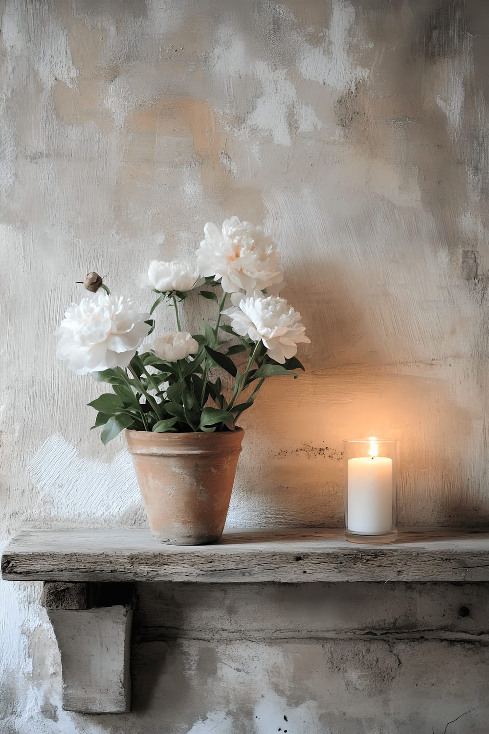 A close-up image of a raw concrete wall which serves as a background for a floating wooden shelf. On the shelf, there's an aged clay pot filled with white peonies. Soft candlelight can be seen reflecting off the shelf and pot.