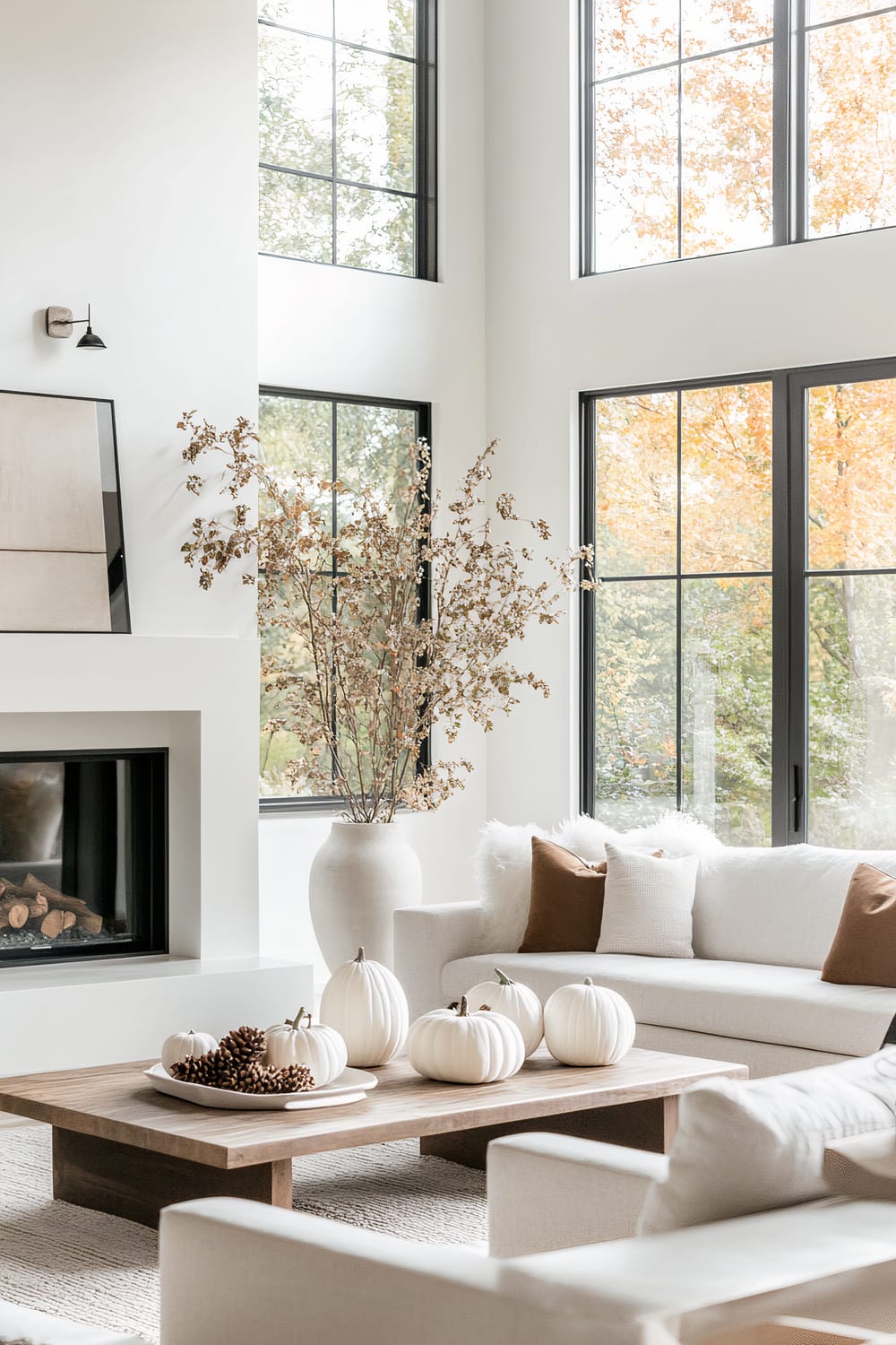 A bright and airy living room with large black-framed windows that let in abundant natural light and offer scenic views of a fall landscape. The room features light-colored furniture, including white sofas accented with brown and white throw pillows. A central wooden coffee table is adorned with white pumpkins and pine cones, contributing to an autumnal theme. To the left, a minimalist fireplace is set into a white wall, with a modern piece of art leaning against the wall above it. A large, white ceramic vase filled with autumnal branches sits next to the fireplace.