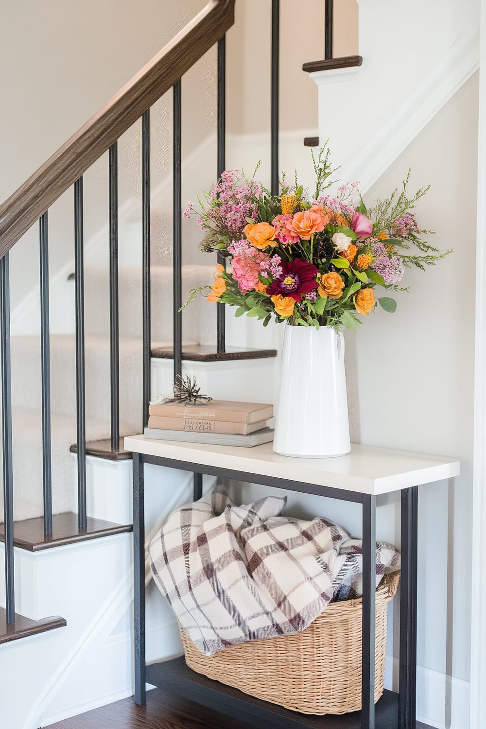 A modern hallway scene featuring a white side table against a staircase. The table is adorned with a white ceramic vase filled with a lively bouquet of mixed flowers in various shades of orange, pink, and purple. Two stacked books with a small decorative item sit next to the vase. Below the table is a woven basket containing a plaid blanket in shades of brown and cream. The staircase has a dark wooden handrail and black vertical balusters, complemented by neutral-toned carpeted steps.