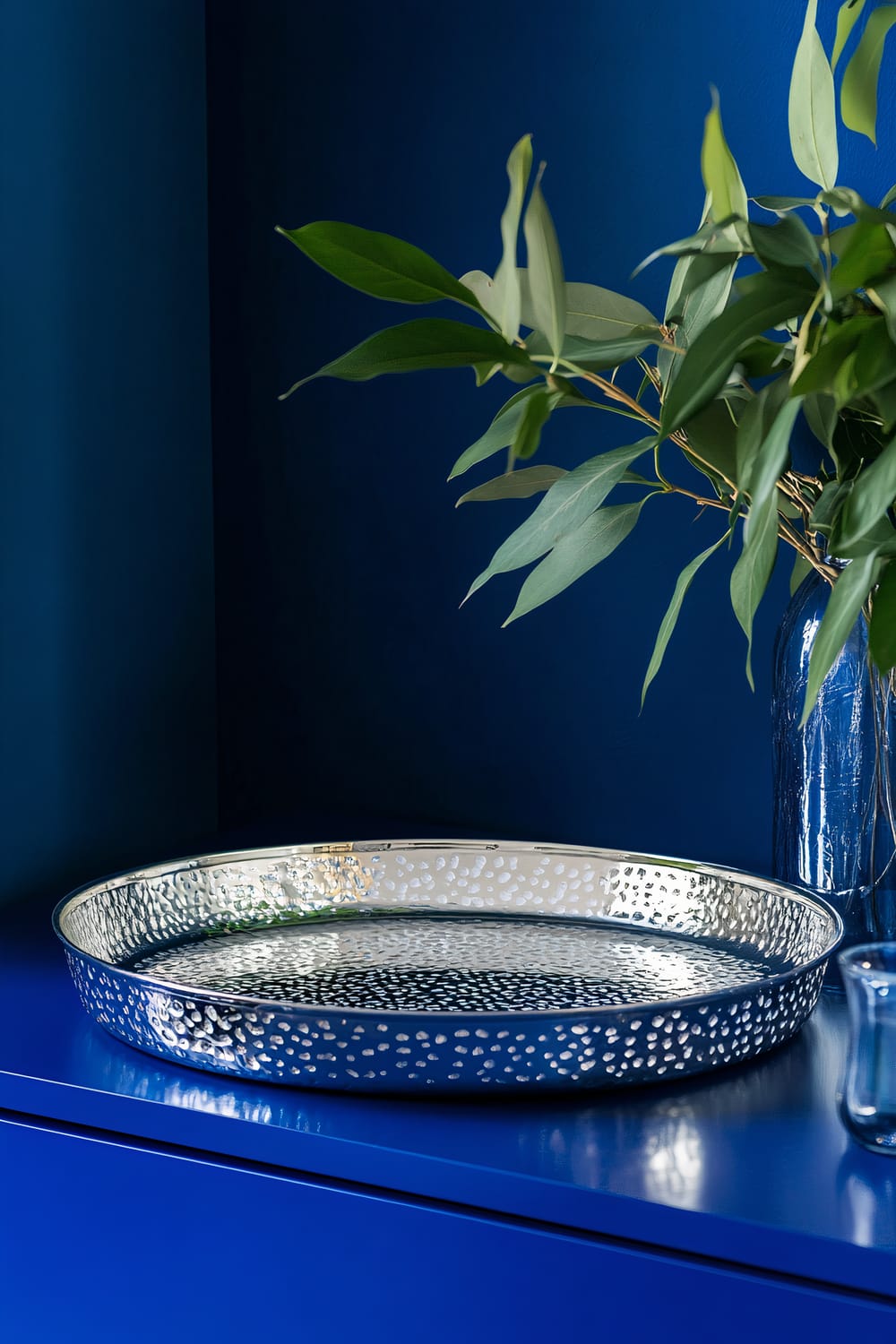 A stylish silver metallic decorative tray with intricate patterns is placed on a vibrant royal blue sideboard. The tray is illuminated by focused lighting, highlighting its reflective surface and detailed design. To the right of the tray is a vase with green leafy branches, and a small glass container is also visible on the sideboard. The background is a deep blue with minimalistic décor, maintaining the focus on the tray.