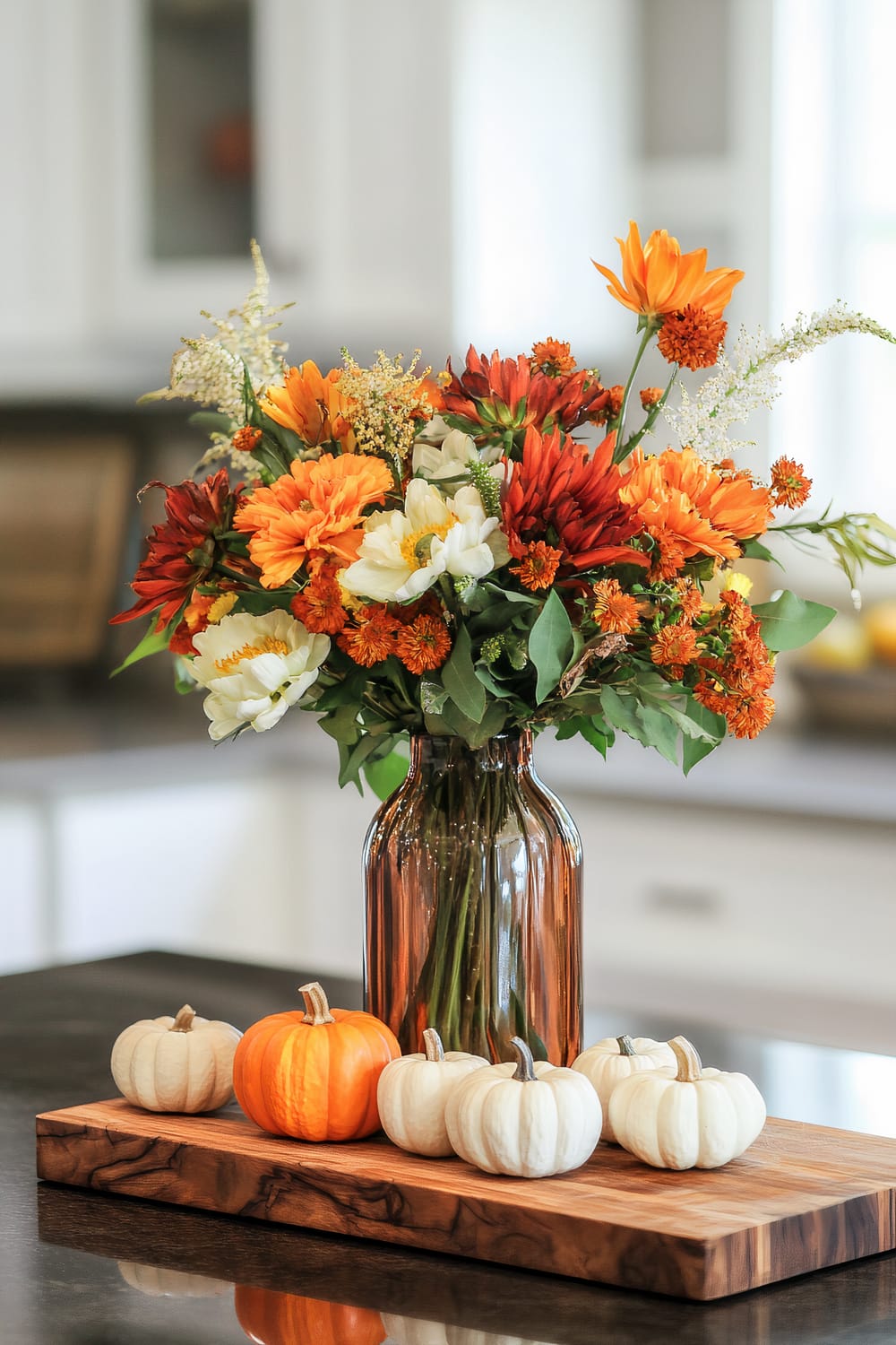 A beautiful arrangement of flowers in autumn hues placed in a glass vase, surrounded by small white and one orange pumpkin, sits atop a wooden cutting board on a kitchen counter. The kitchen background features white cabinetry and a neutral color scheme.