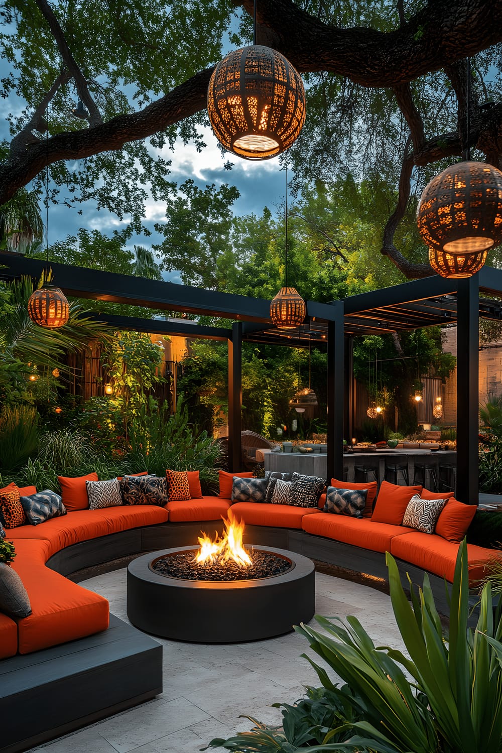 A contemporary evening garden lounge with a circular, black metal-framed fire pit at the center. Vibrantly colored, modular outdoor sofas in orange and charcoal gray are arranged around the fire pit. Large outdoor cushions adorned with geometric designs and colorful lanterns hanging from nearby trees add to the ambiance. All elements are illuminated by the warm firelight and soft ambient lighting, creating an inviting outdoor space.